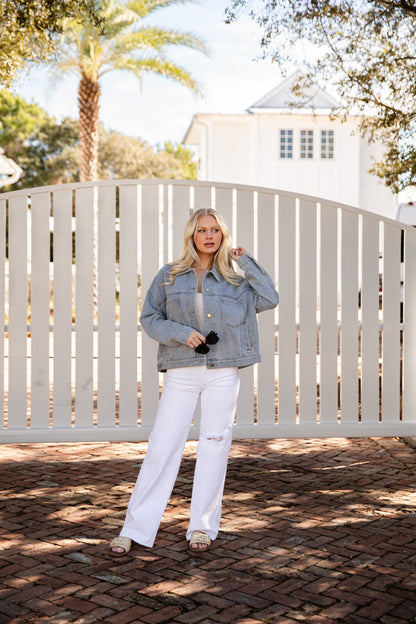 Woman in denim jacket and white pants standing in front of a white fence with trees in the background