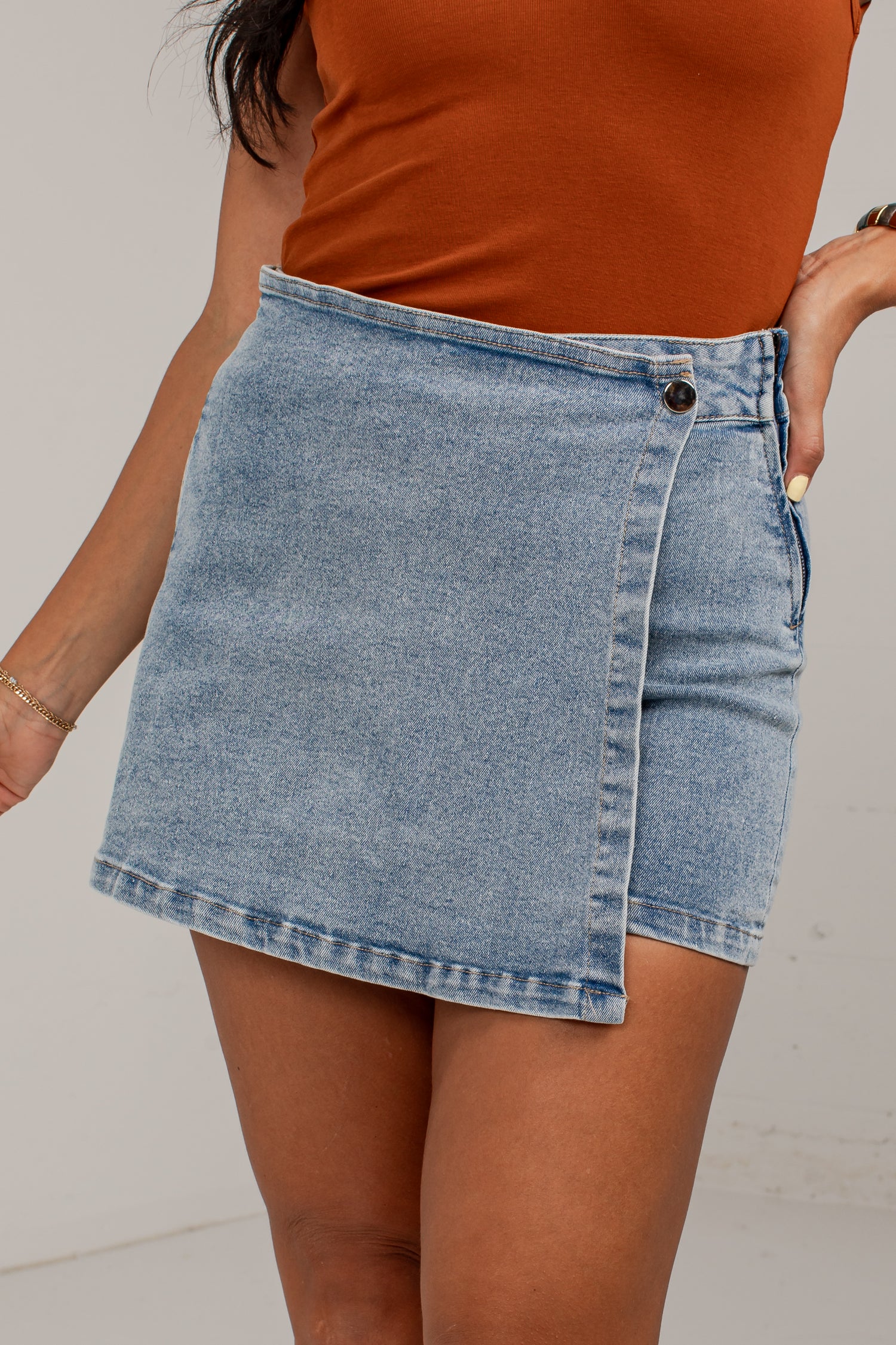 A person wearing a light-wash denim wrap mini skirt and an orange-brown ribbed top, shown from the chest down against a plain light background.