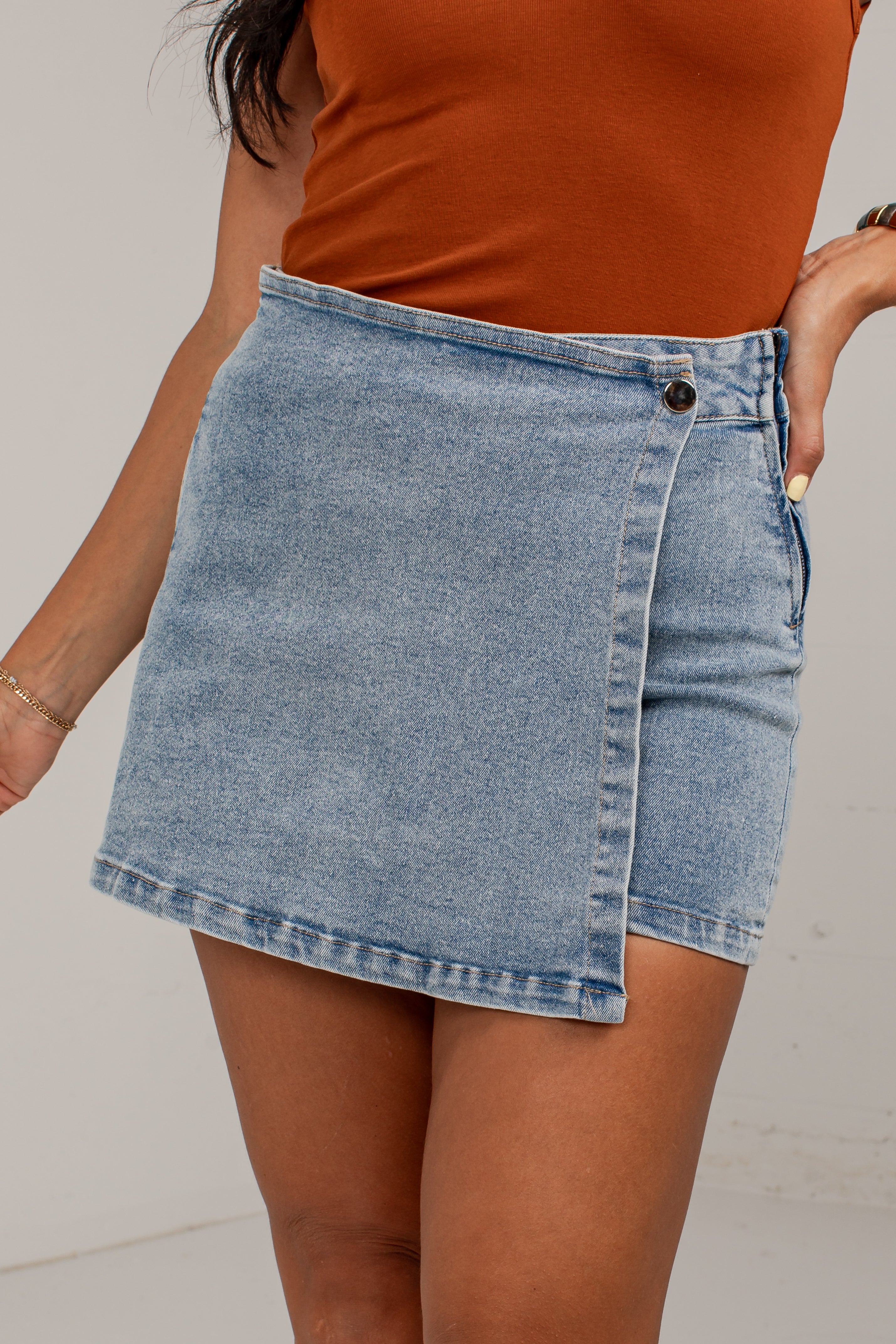 A person wearing a light-wash denim wrap mini skirt and an orange-brown ribbed top, shown from the chest down against a plain light background.