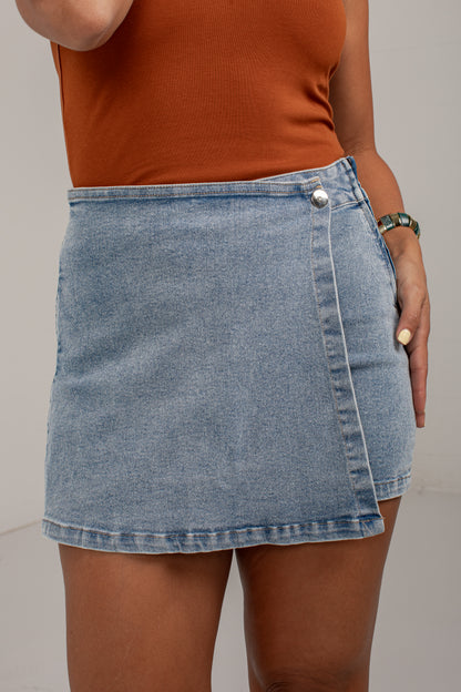 A person wearing a light-wash blue denim mini skirt with a diagonal front zipper and a silver-tone snap button, paired with an orange-brown ribbed tank top against a plain light gray studio background.