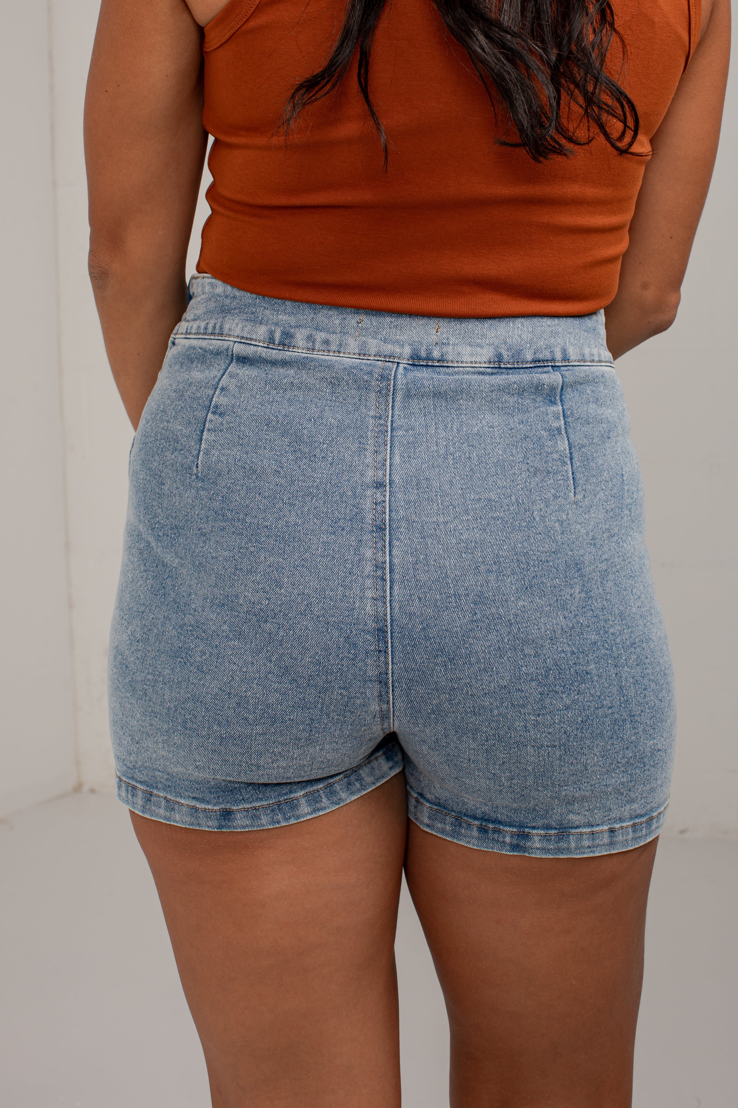 Rear view of a person wearing light-wash blue denim shorts and an orange sleeveless top against a plain light background.