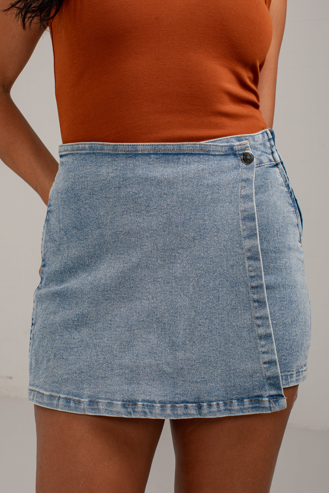 A light blue denim wrap mini skirt worn by a model, paired with a ribbed orange-brown tank top, shown from the chest down against a neutral light gray background.