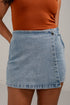 A light blue denim wrap mini skirt worn by a model, paired with a ribbed orange-brown tank top, shown from the chest down against a neutral light gray background.