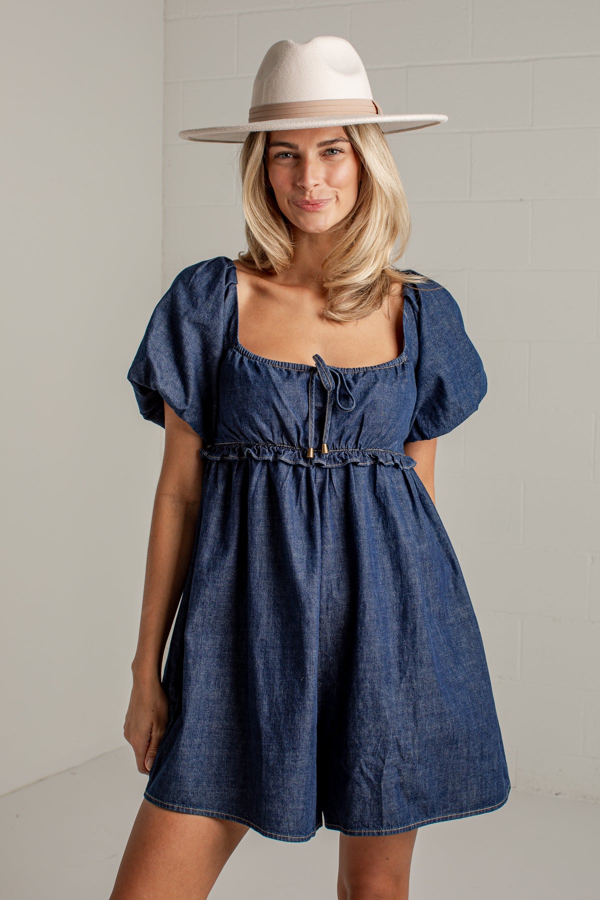 A woman wearing a blue denim short romper with puff sleeves and a square neckline, styled with a wide-brimmed cream hat, standing against a white brick wall.