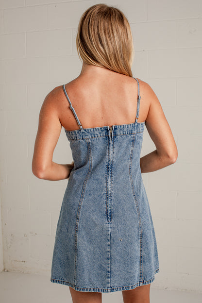 A woman viewed from behind wearing a light blue denim mini dress with thin spaghetti straps and a buttoned back, standing against a white painted brick wall.