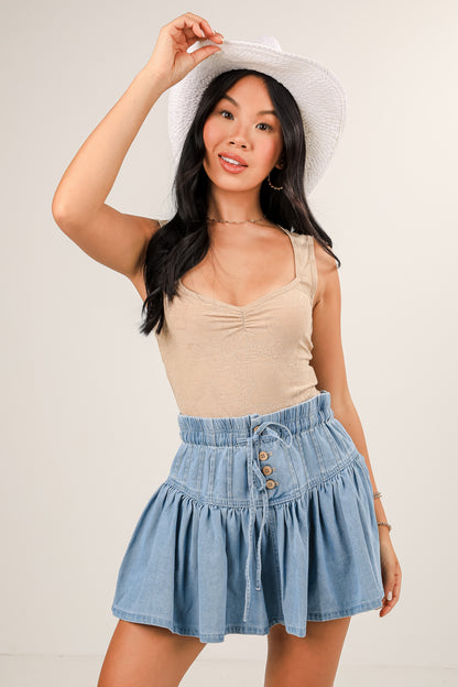 Woman wearing a beige tank top and denim skirt with a white hat on a plain background