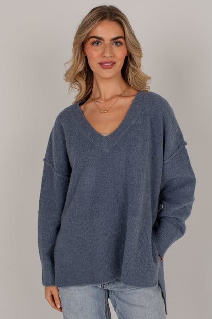 A woman wearing an oversized, heathered slate blue V-neck knit sweater with light-wash jeans, standing against a neutral light gray studio background.