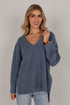 A woman wearing a heathered slate blue V-neck knit sweater and light-wash jeans, standing against a neutral light gray studio background.