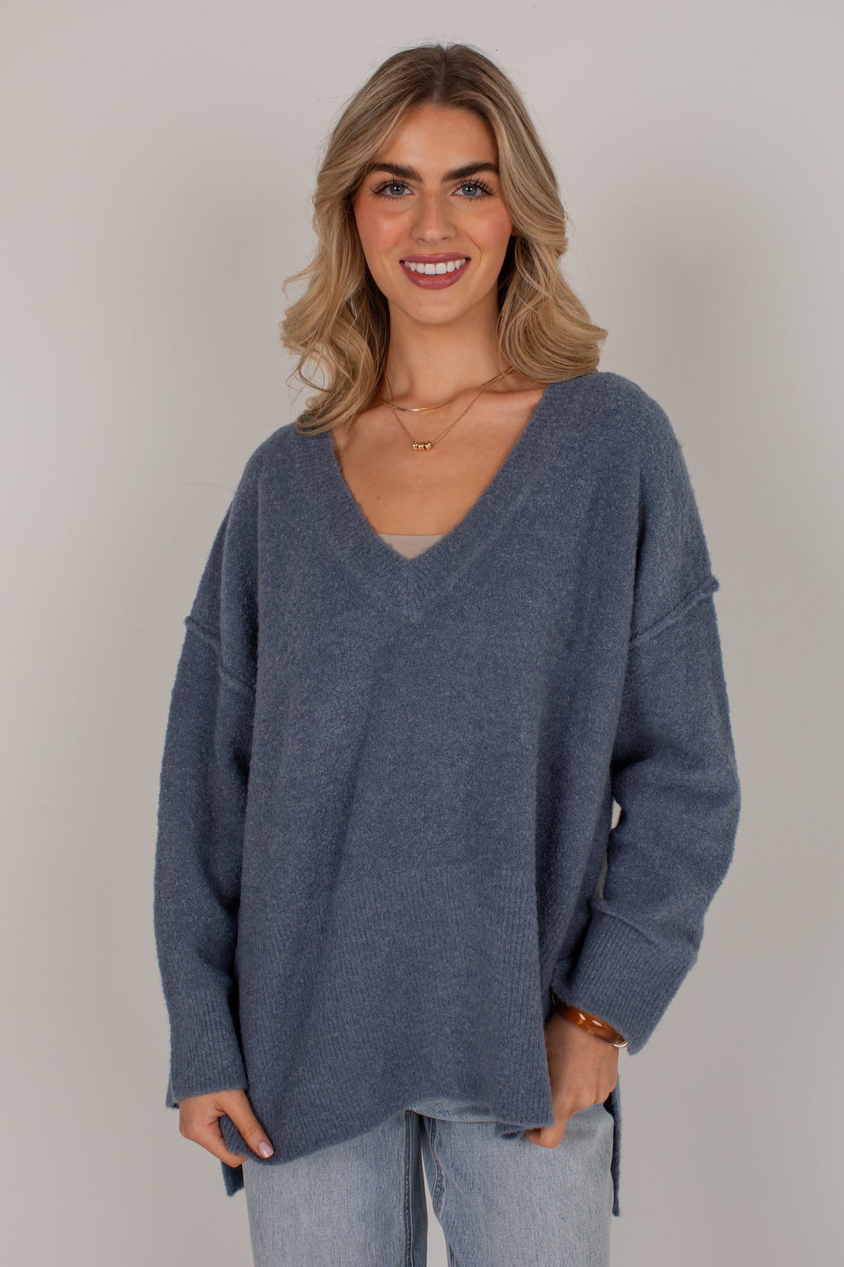 A woman wearing an oversized, long-sleeve V-neck knit sweater in a dusty slate blue color, paired with light-wash jeans, against a plain light gray studio background.