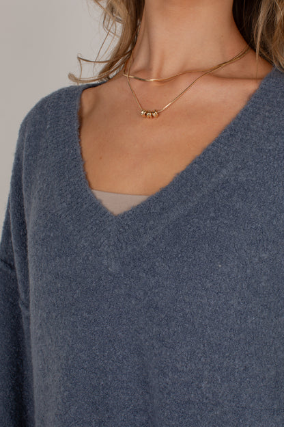 Close-up of a person wearing a slate blue V-neck sweater and layered gold necklaces, including a delicate chain and a beaded pendant.