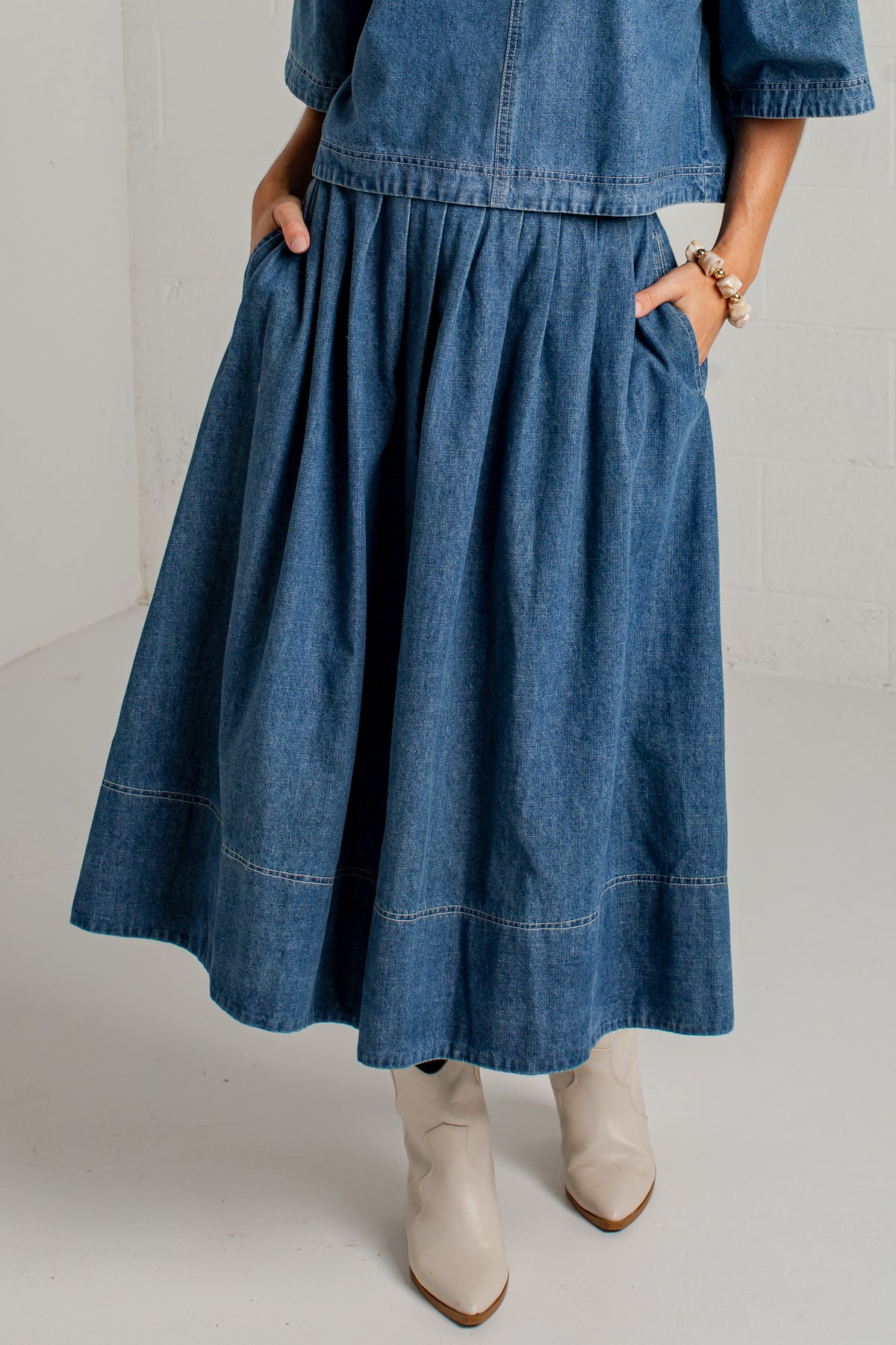 A person wearing a blue denim midi skirt with side pockets, paired with a matching denim top and beige ankle boots, shown from the waist down against a light studio background.