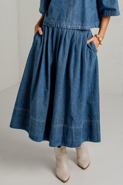 A person wearing a blue denim midi skirt with side pockets, paired with a matching denim top and beige ankle boots, shown from the waist down against a light studio background.