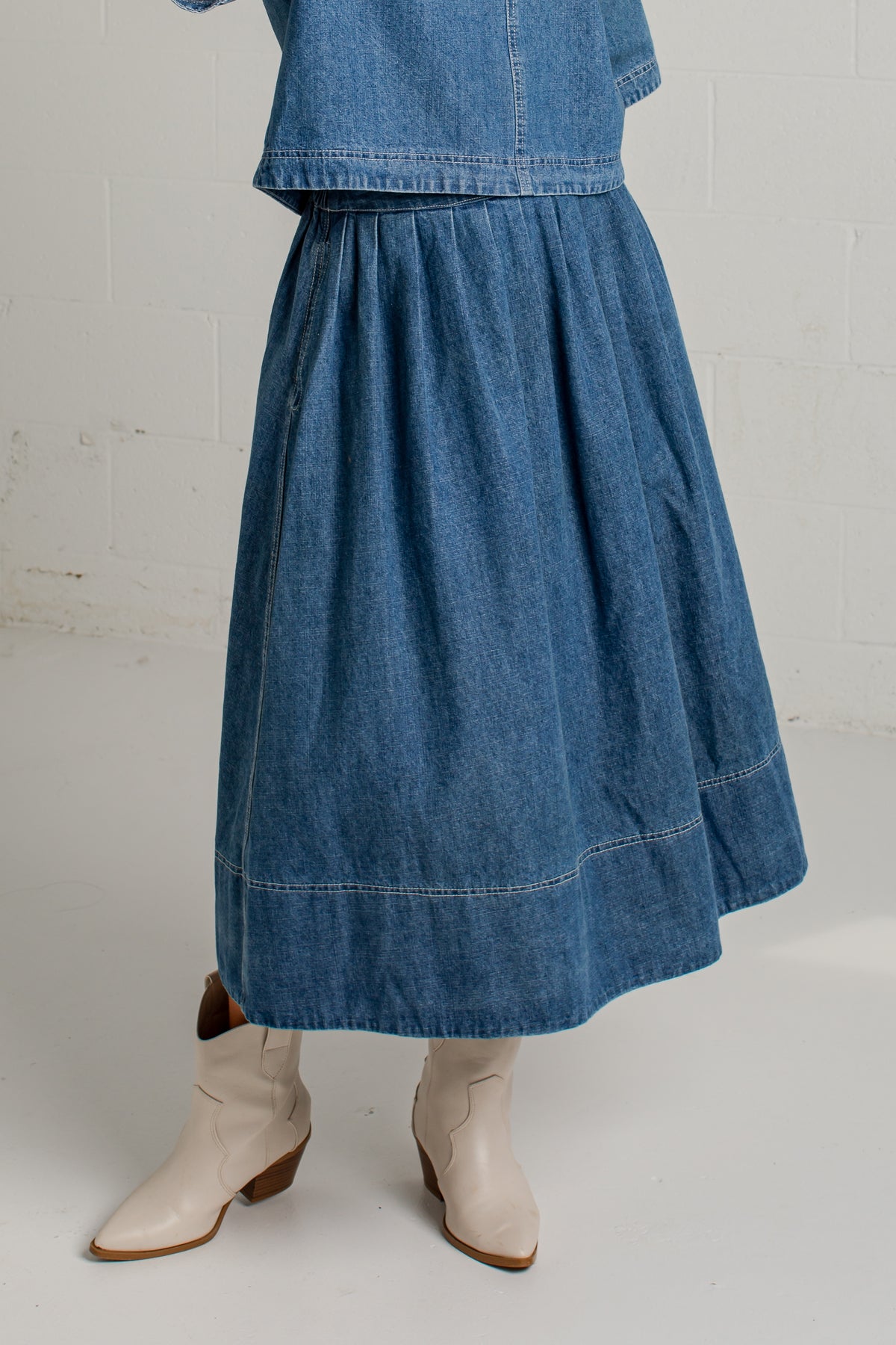 A person wearing a blue denim midi skirt with a matching cropped denim jacket and light beige cowboy boots, standing against a white brick wall.