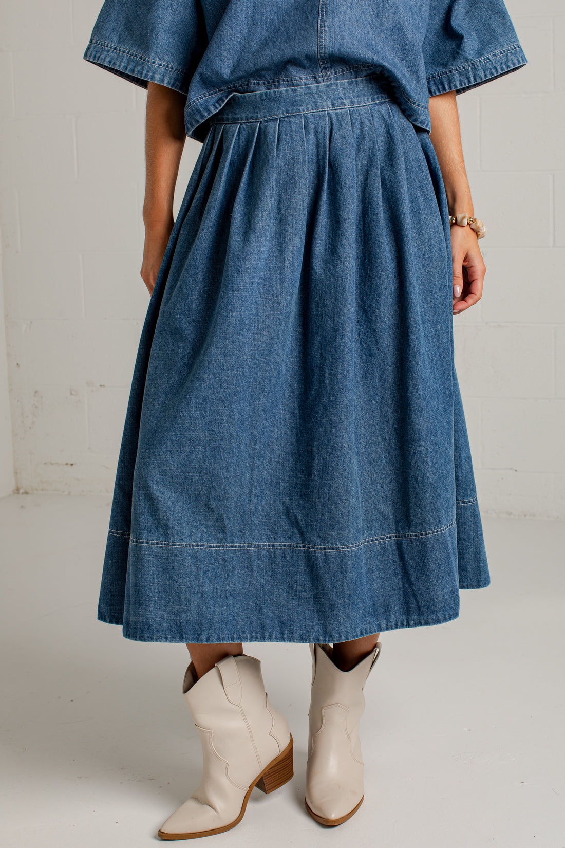 A person wearing a blue denim midi skirt with a pleated, gathered waist and hem, paired with cream-colored western-style ankle boots, shown from the waist down against a white studio wall.