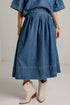 A person wearing a blue denim midi skirt with a pleated, gathered waist and hem, paired with cream-colored western-style ankle boots, shown from the waist down against a white studio wall.