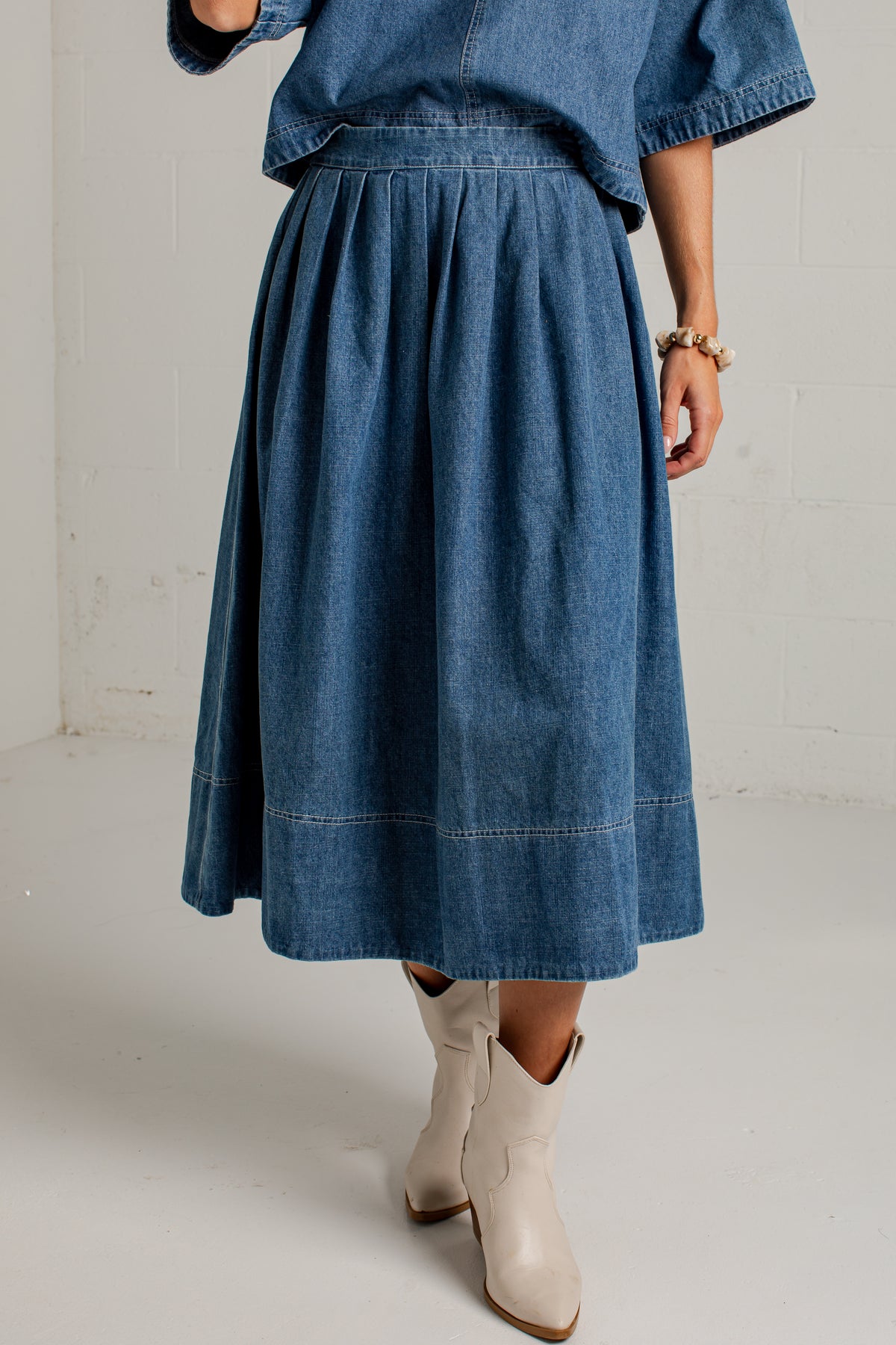 A person wearing a blue denim midi skirt with a matching denim top and off-white cowboy boots, standing against a white brick wall.