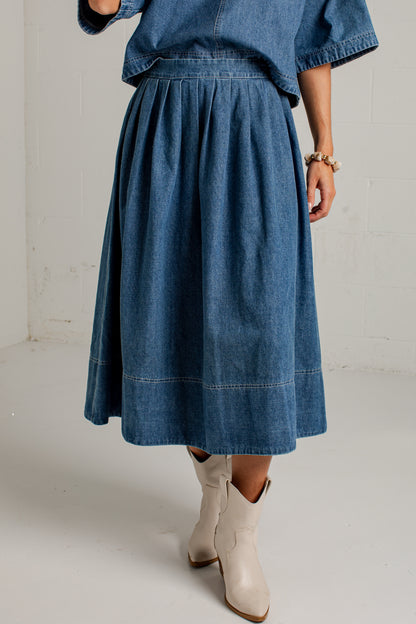 A person wearing a blue denim midi skirt with a matching denim top and off-white cowboy boots, standing against a white brick wall.