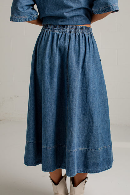 A person wearing a blue denim midi skirt with an elastic waistband and light beige cowboy boots, shown from the back against a white studio wall.