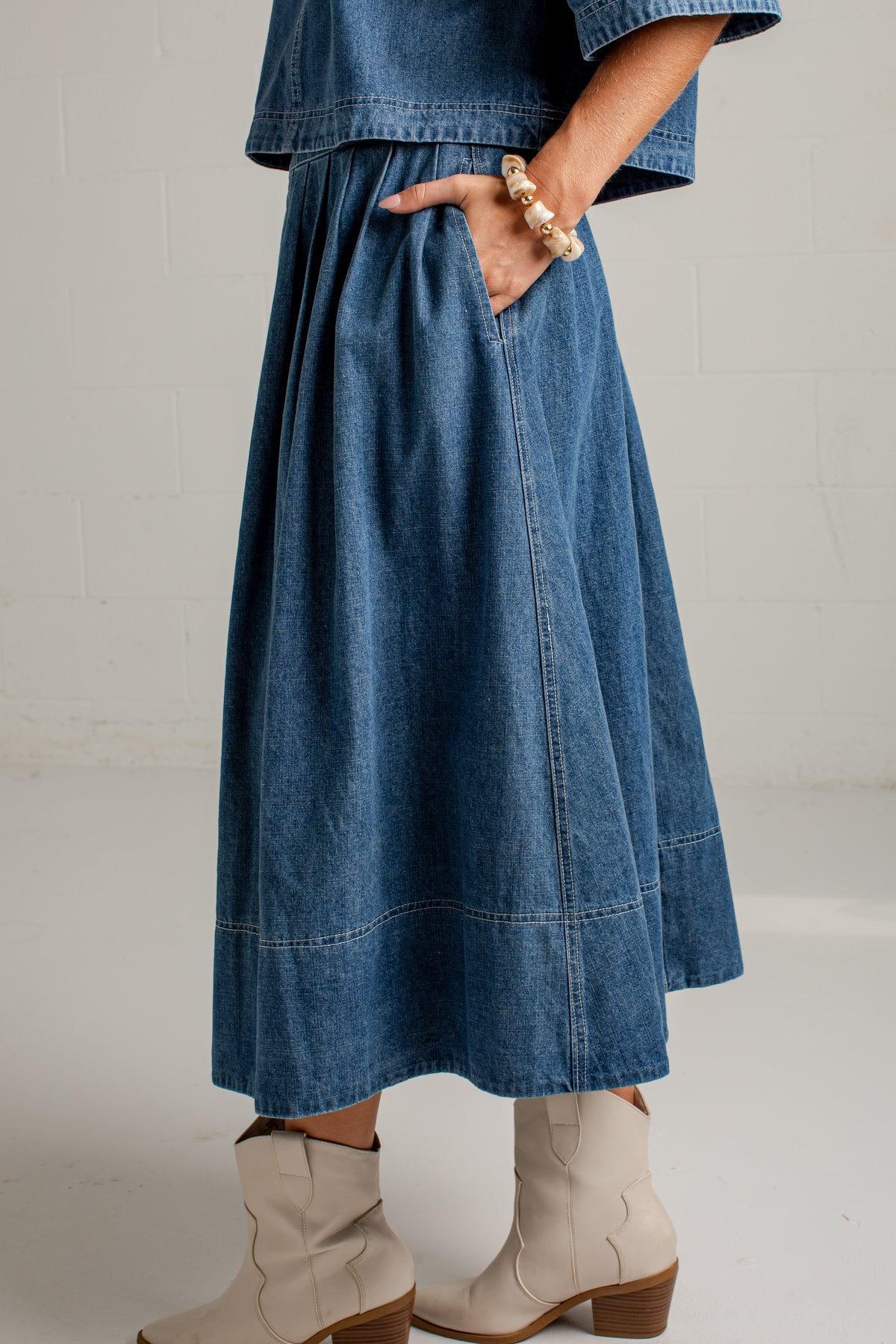 A person wearing a blue denim midi skirt with side pockets, paired with a matching denim top and beige ankle boots, standing against a white brick wall.