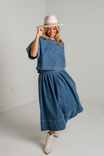 A woman modeling a matching blue denim two-piece set consisting of a short-sleeve cropped top and a high-waisted pleated midi skirt, styled with a beige wide-brim hat and beige ankle boots against a white brick wall background.