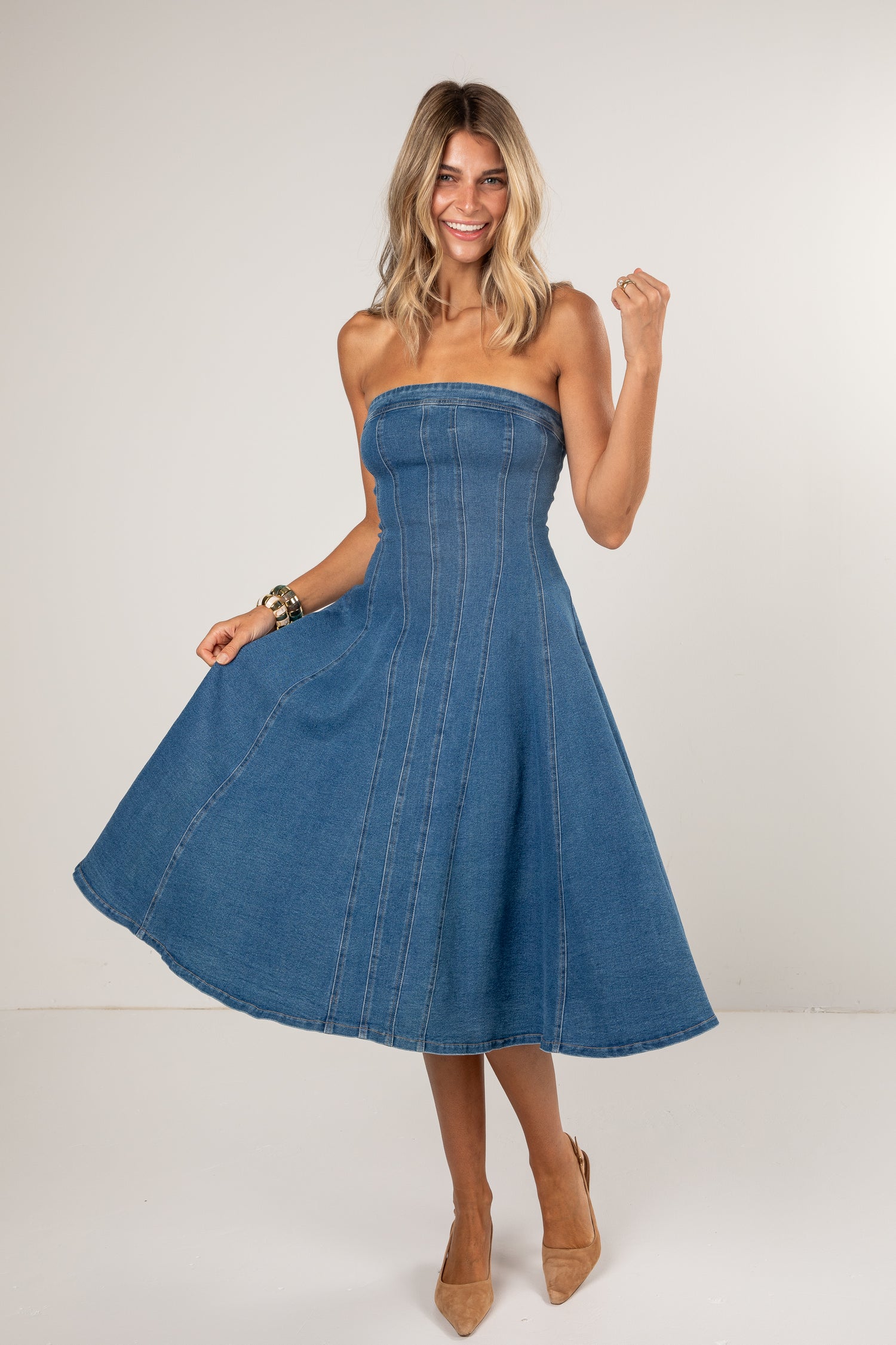 A woman modeling a strapless blue denim midi dress with a flared skirt, paired with beige heels, against a plain light gray studio background.