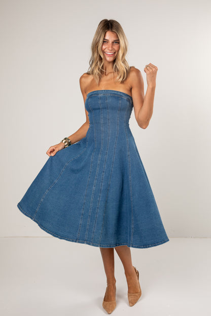 A woman modeling a strapless blue denim midi dress with a flared skirt, paired with beige heels, against a plain light gray studio background.