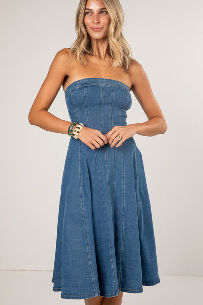 A woman modeling a strapless blue denim dress with a fitted corset-style bodice and a flared skirt, photographed against a plain light studio background.