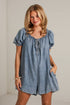 A woman modeling a light blue denim romper with puff sleeves, a smocked neckline, and front tie details, standing against a white brick wall.