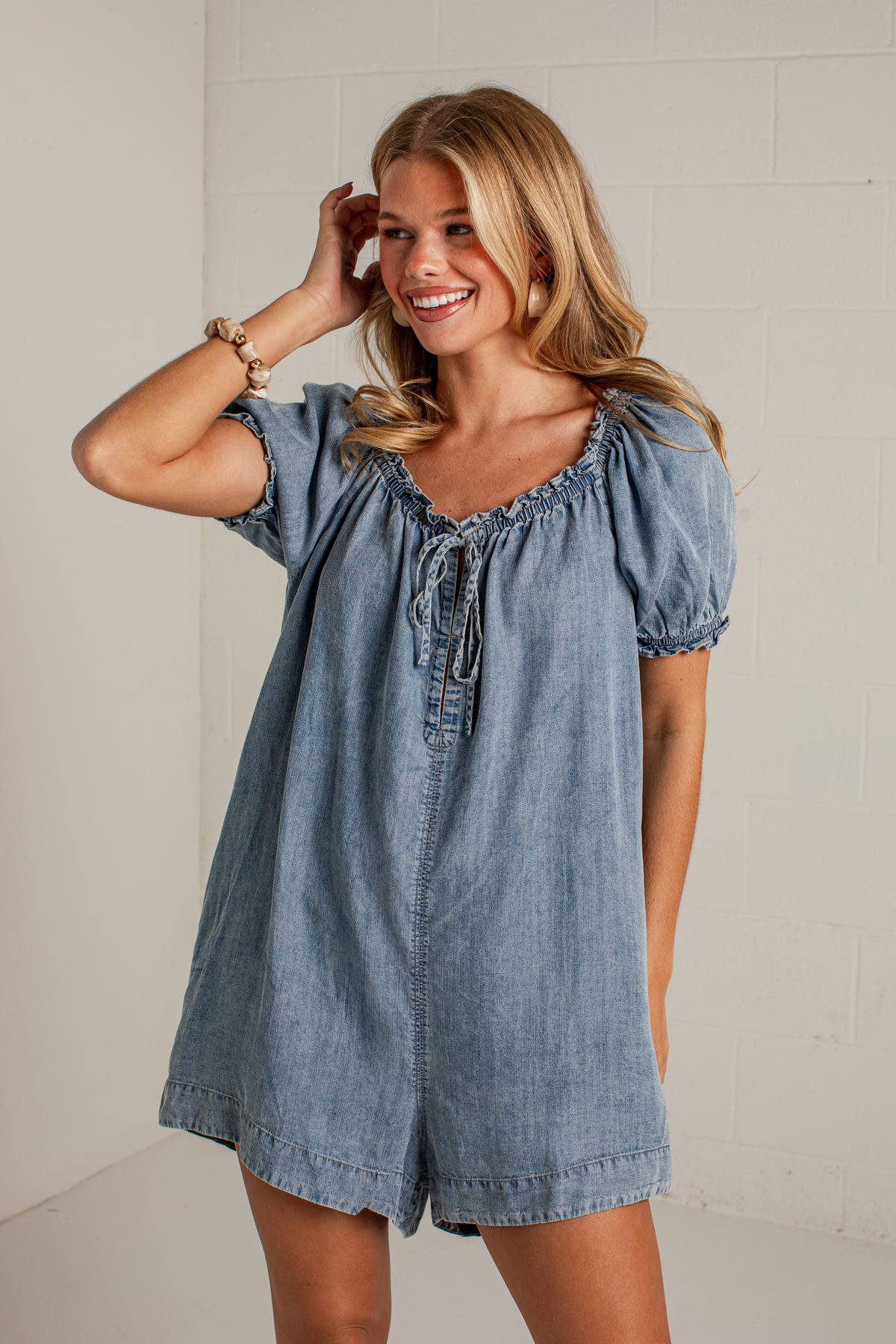 A woman modeling a light blue denim short romper with puff sleeves and a tie neckline, standing against a white brick wall.