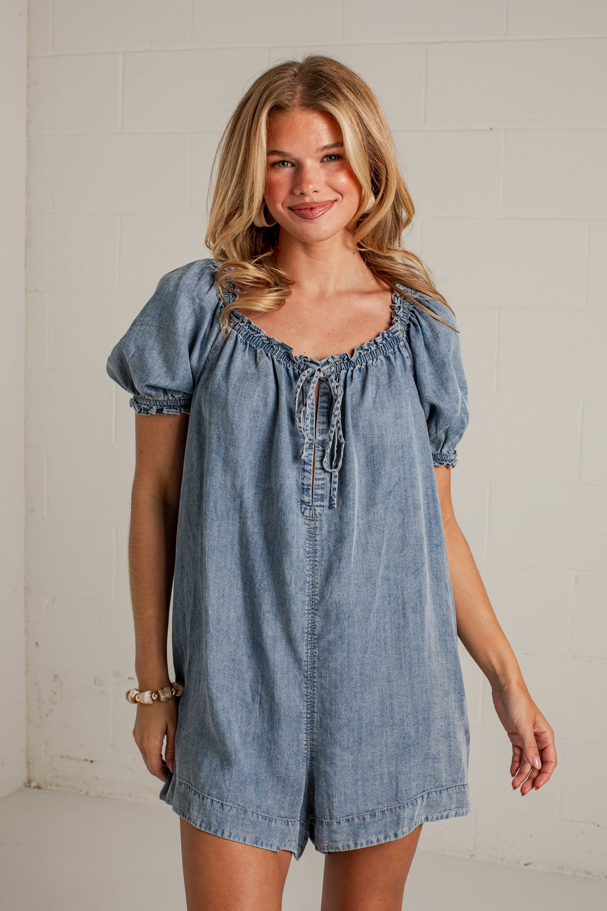 A woman modeling a light blue denim romper with puff sleeves, a smocked neckline, and a front tie closure, standing against a white brick wall.