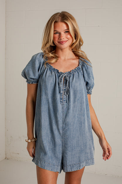 A woman modeling a light blue denim romper with puff sleeves, a smocked neckline, and a front tie closure, standing against a white brick wall.