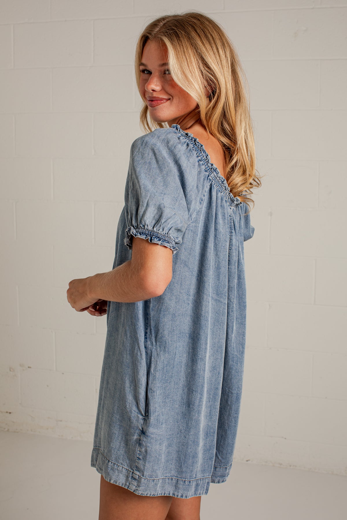 A woman wearing a light blue denim short-sleeve dress with ruffled trim, photographed against a white brick wall.
