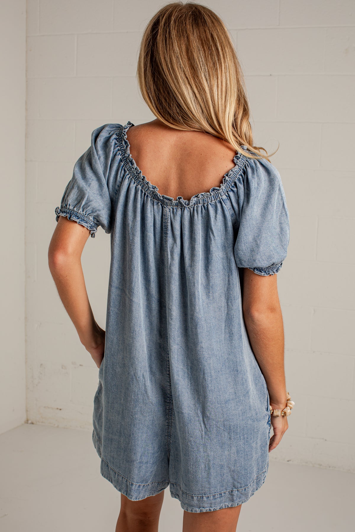 A woman viewed from behind wearing a light blue denim romper with puff sleeves and a ruffled scoop neckline, standing against a white brick wall.