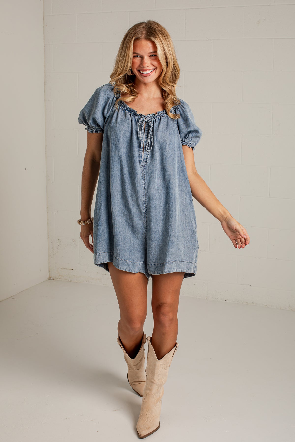 A woman modeling a light blue denim short-sleeve tunic dress with puff sleeves and a tie neckline, paired with beige cowboy boots, in a bright studio setting.