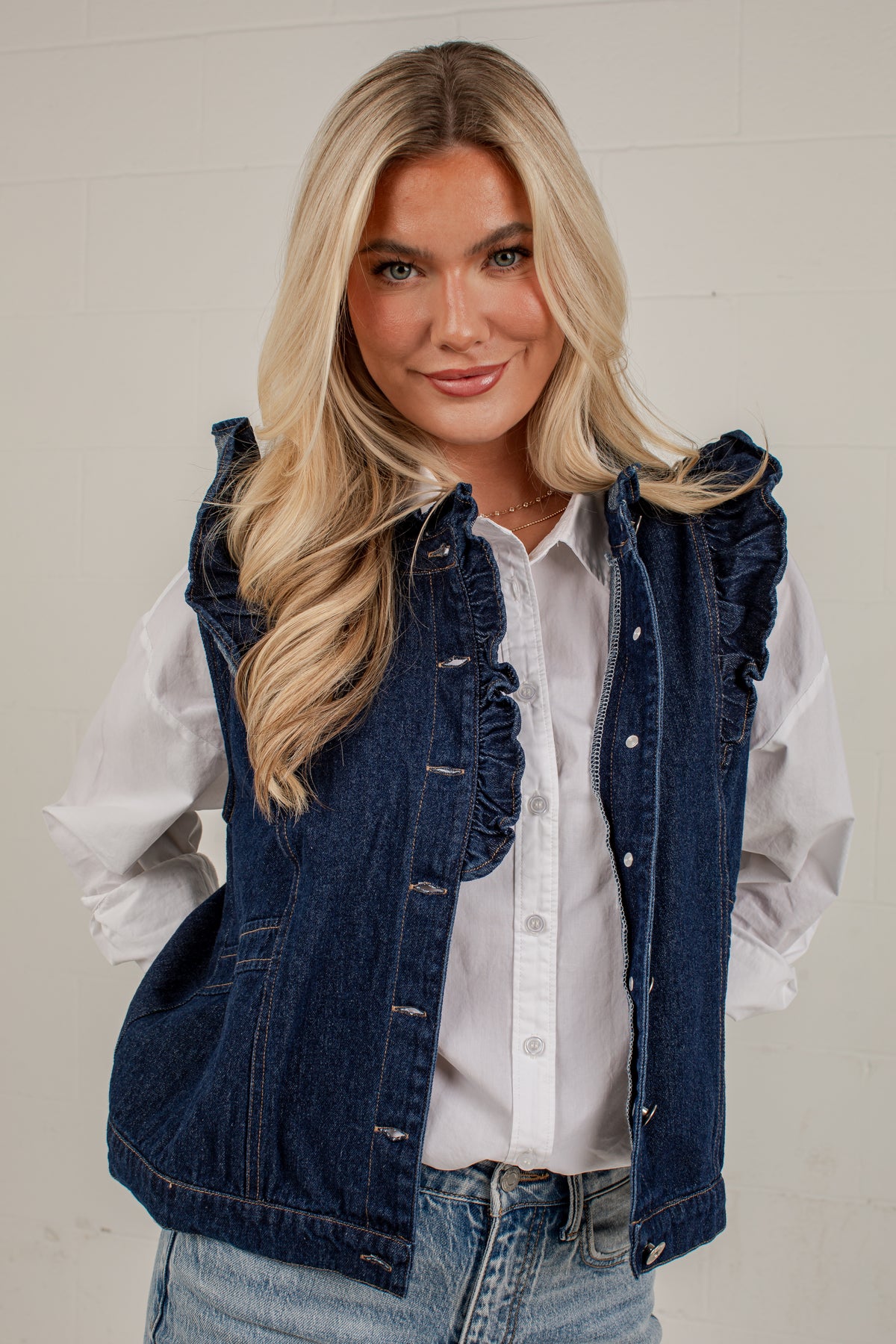 A blonde woman wearing a dark blue denim vest with ruffle trim over a white button-up shirt and light-wash jeans, standing against a white paneled wall.