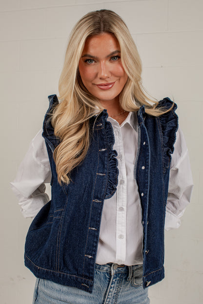 A blonde woman wearing a dark blue denim vest with ruffle trim over a white button-up shirt and light-wash jeans, standing against a white paneled wall.