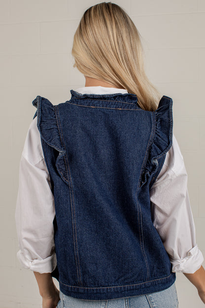 A woman viewed from behind wearing a dark blue denim vest with ruffle details on the shoulders over a white long-sleeve shirt and light blue jeans.