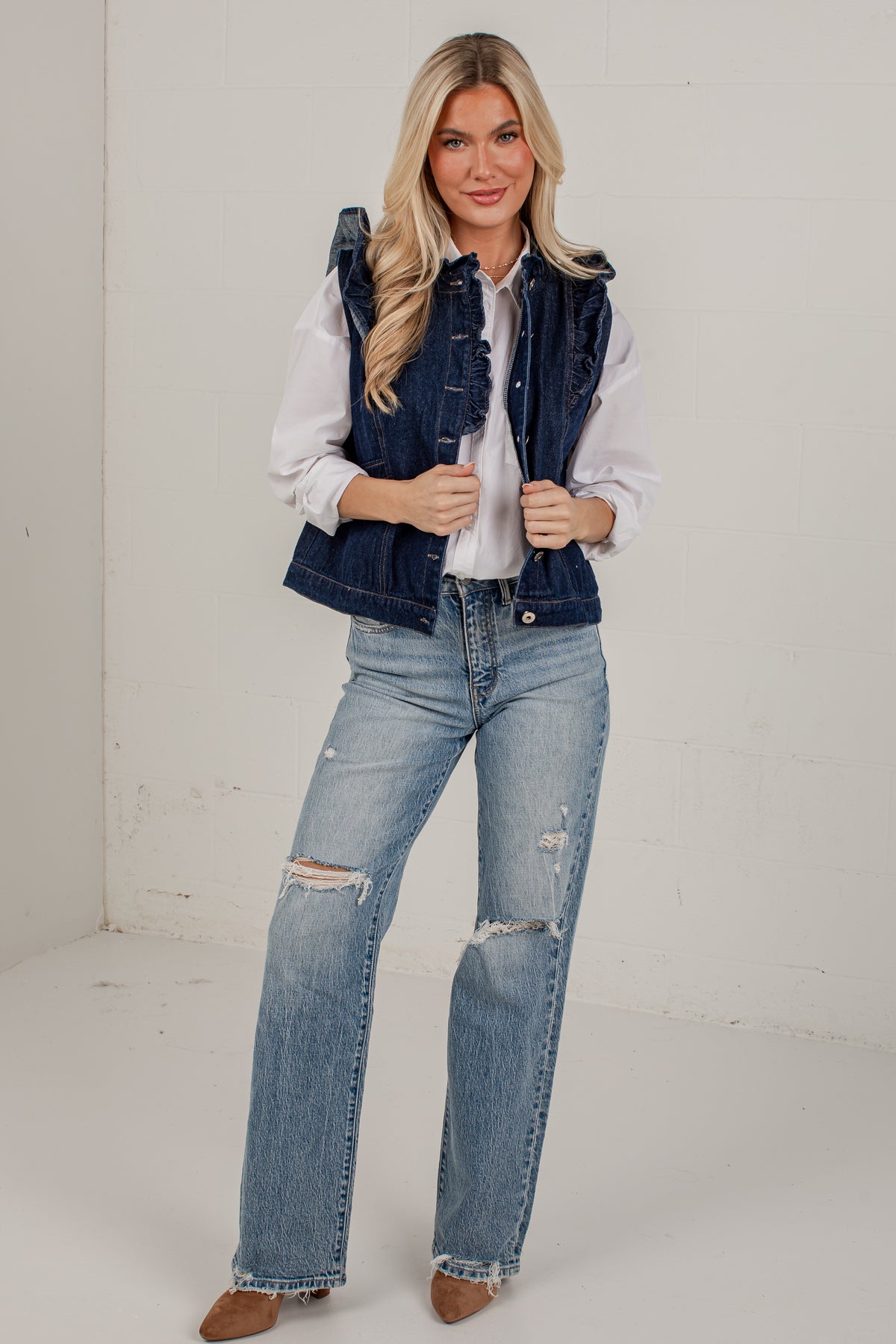 A woman modeling a dark wash denim vest over a white button-down shirt and light-wash distressed jeans, standing against a plain off-white wall.