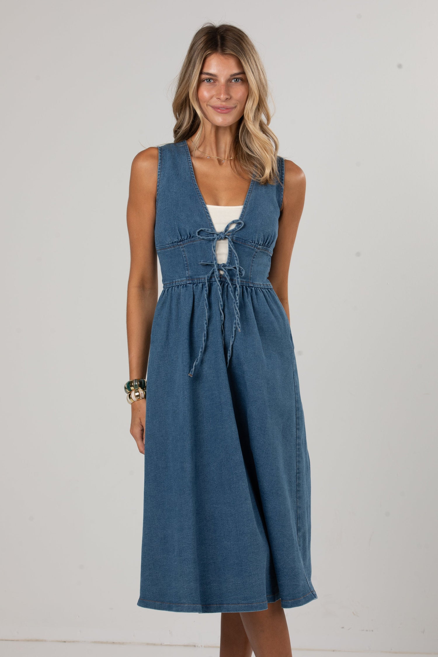 A woman modeling a sleeveless blue denim midi dress with a tie-front detail over a white top, standing against a plain light gray studio background.