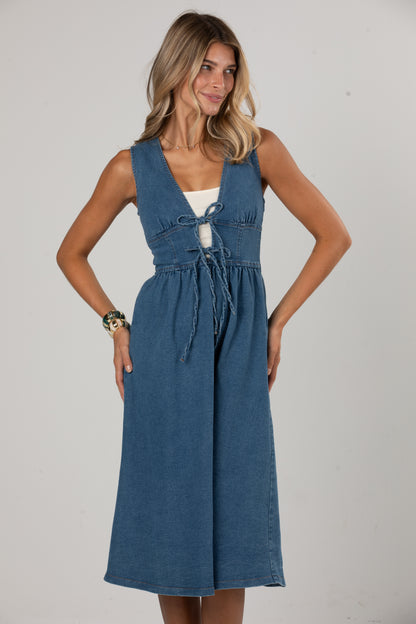 A woman modeling a sleeveless blue denim midi dress with a tie-front detail over a white underlayer, photographed against a plain light gray studio background.