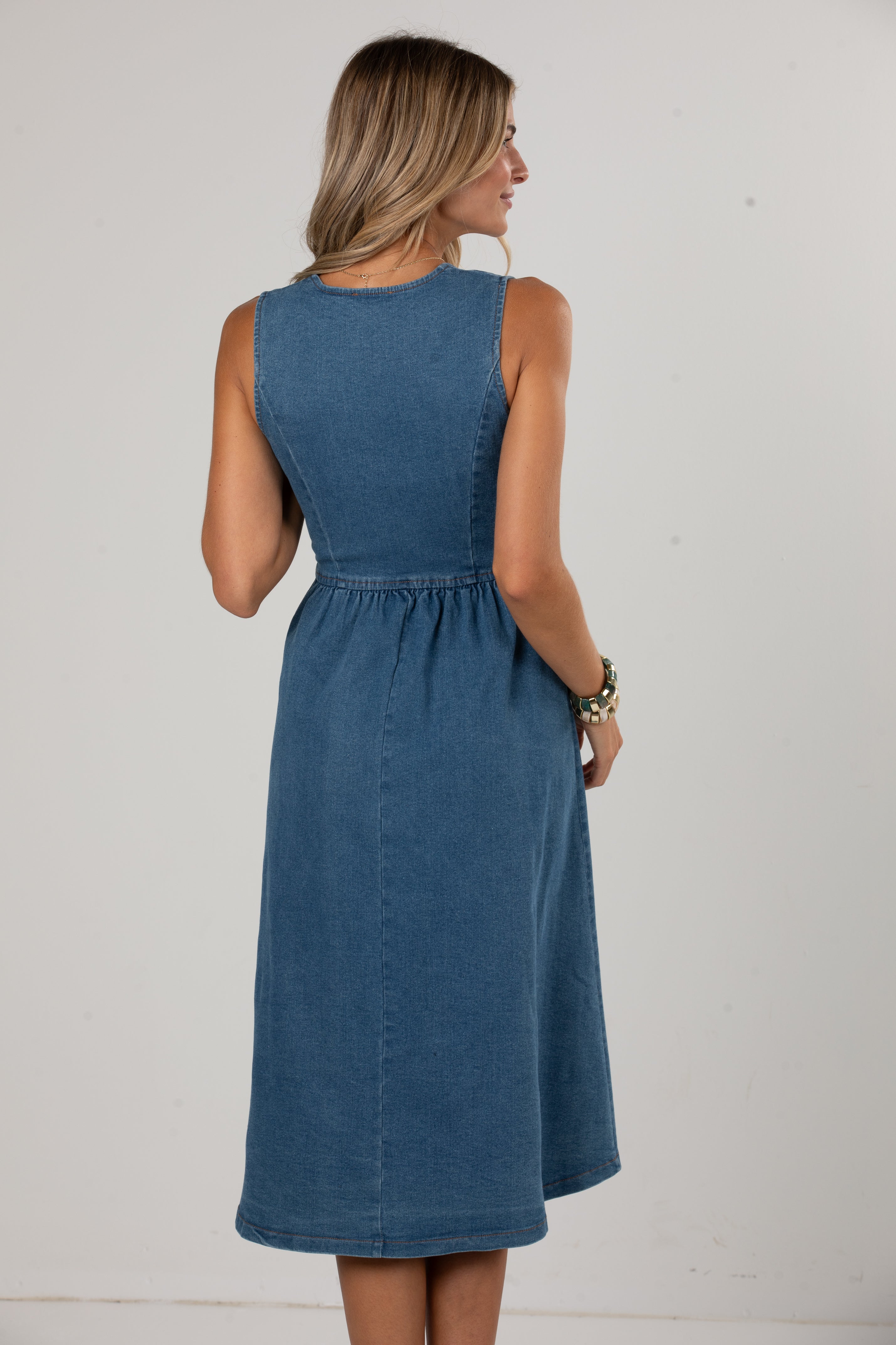 A woman wearing a sleeveless blue denim midi dress, shown from the back against a plain off-white studio background.