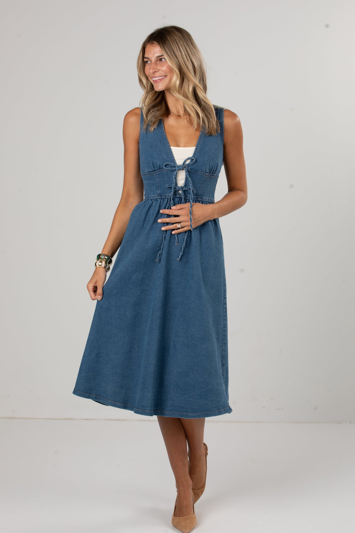 A woman modeling a sleeveless blue midi dress with a tie-front detail and a white underlayer visible at the neckline, standing against a plain light gray studio background.