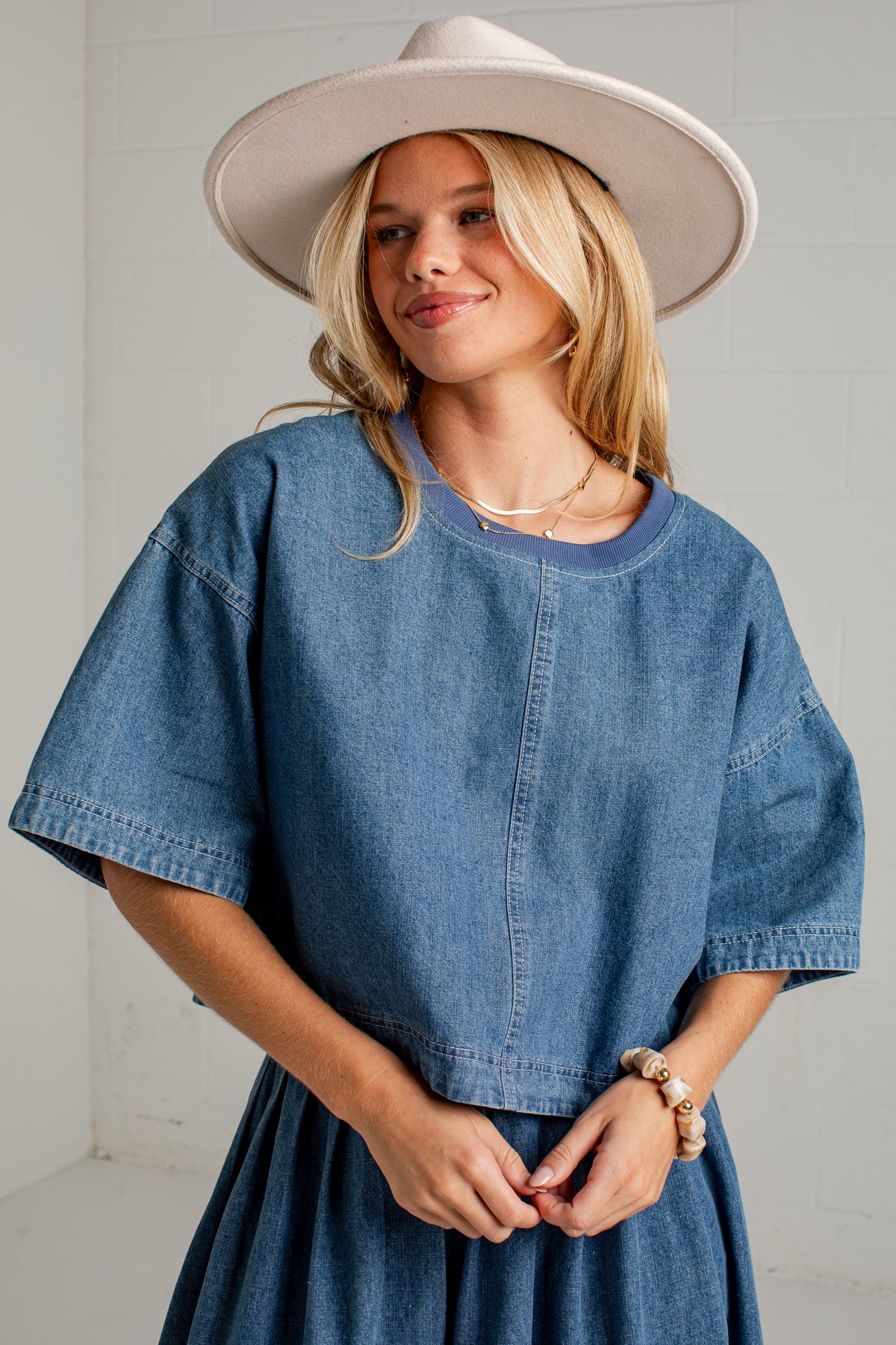 A woman wearing a loose-fitting blue denim dress with wide short sleeves and a matching skirt, styled with a wide-brimmed cream hat and layered gold necklaces.