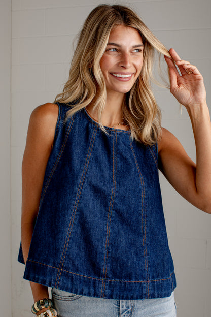 A woman wearing a sleeveless dark blue denim top with orange contrast stitching, paired with light-wash jeans, posing against a light gray paneled wall.