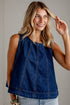 A woman wearing a sleeveless dark blue denim top with orange contrast stitching, paired with light-wash jeans, posing against a light gray paneled wall.
