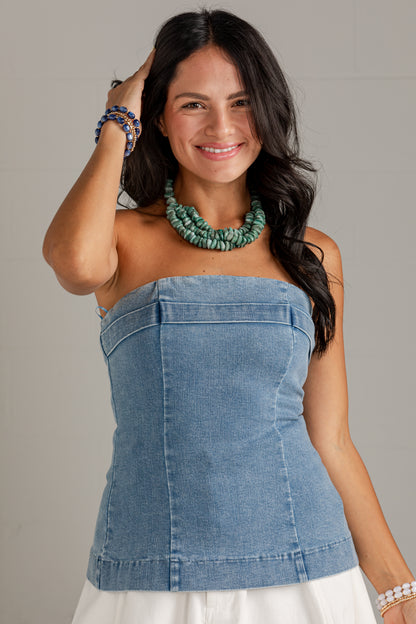A woman wearing a light blue denim strapless tube top with a corset-style front panel, paired with a white skirt and a chunky green beaded necklace, posing against a neutral light gray background.