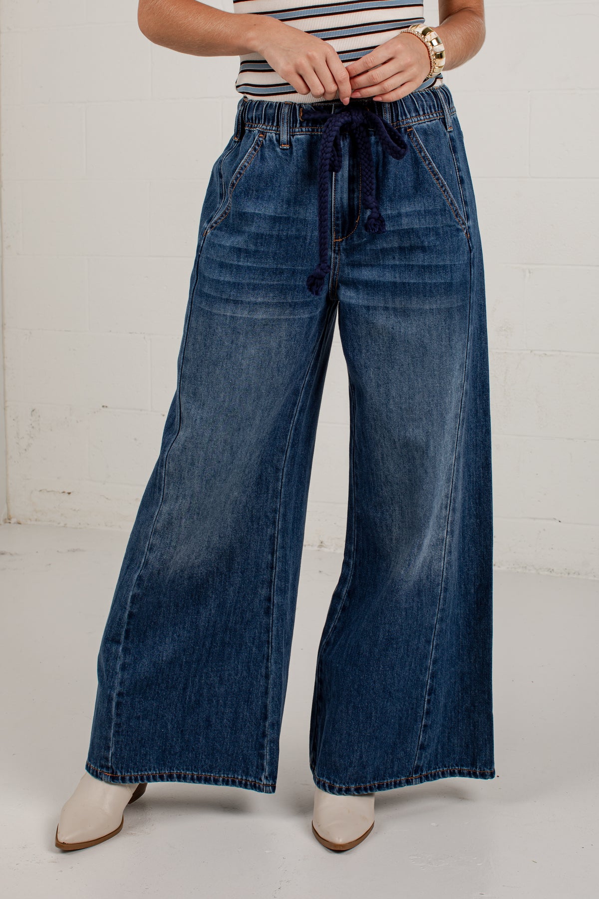 A person wearing wide-leg blue denim jeans with a dark blue drawstring waist, paired with a striped top and white pointed-toe heels, standing against a white wall.