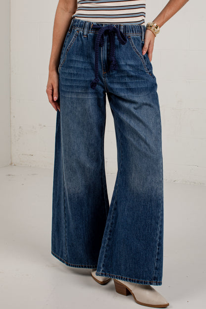 A person wearing wide-leg blue denim jeans with a dark blue drawstring waistband, paired with a striped top and beige heeled shoes, standing against a white wall.