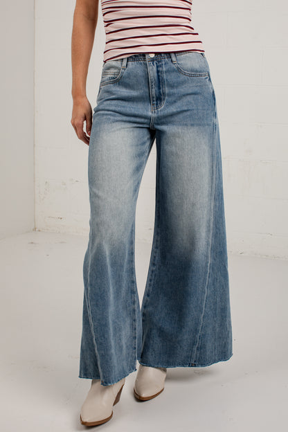 A person wearing wide-leg blue denim jeans with a faded wash, paired with a striped top and light-colored heeled boots, standing against a plain light gray wall.