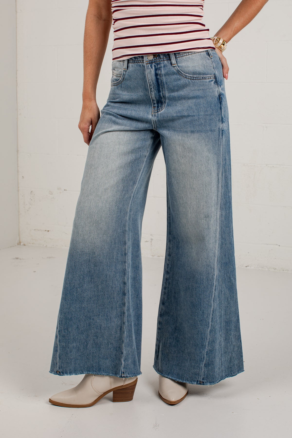 A model wearing a pair of wide-leg blue denim jeans with a faded wash, paired with a red and white striped top and beige heeled boots, standing against a white brick wall.